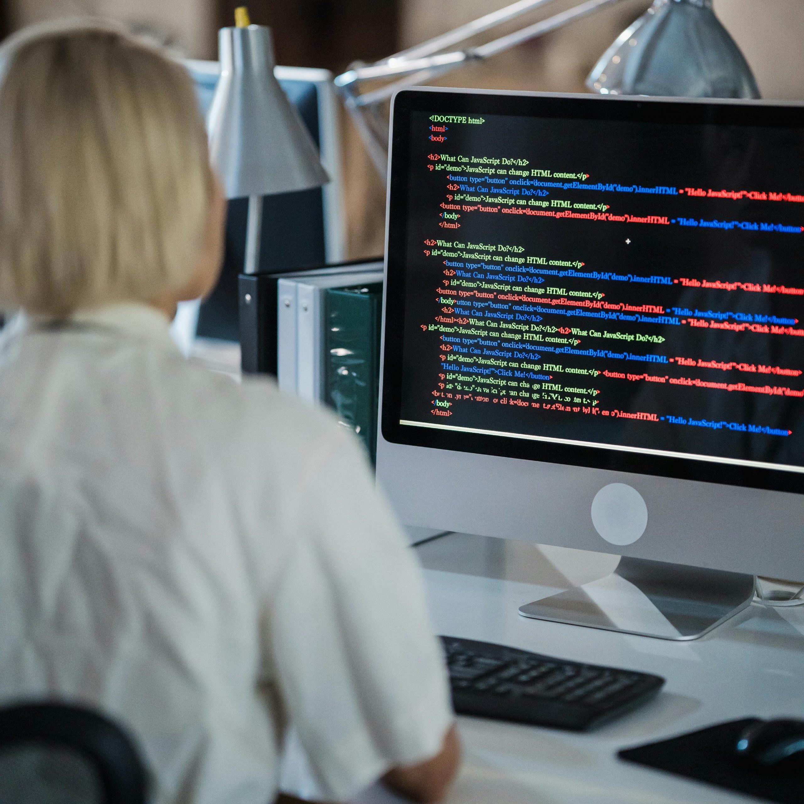 Professional woman coding at office desk with two monitors and desk lamps.